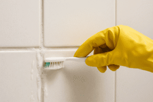Hand in yellow glove scrubbing bathroom grout with toothbrush and natural paste—how to clean grout without chemicals.